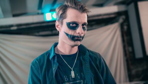 Portrait d'Halloween d'un homme accoudé à un bar, avec un maquillage sombre autour des yeux et une cicatrice factice partant des lèvres. Il porte une chemise ouverte sur un t-shirt avec un pendentif. Son expression est sérieuse. Une pinte de bière est posée à côté de lui.