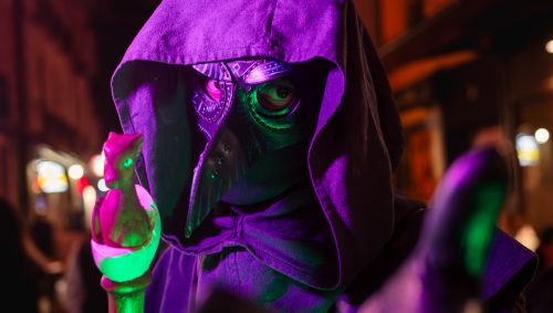Déguisement d'Halloween d'un médecin de la peste, portant une cape à capuche violette et un masque avec un long bec, tendant la main vers la caméra tout en tenant un bâton vert surmonté d'un petit dragon violet.