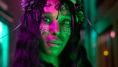 Portrait d'une jeune femme arborant un maquillage d'Halloween sur le visage, notamment des motifs en forme de toiles d'araignées blanches sous les yeux, et portant une couronne de fleurs sombres, le tout éclairé par des leds roses et verts.