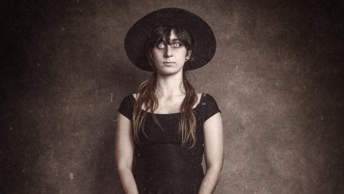 Portrait d'une jeune femme déguisée en sorcière pour Halloween, regard fixe et yeux blancs, portant un grand chapeau noir, une robe noire et des bas rayés violets et noirs, le tout dans un style vintage sur fond marron foncé.