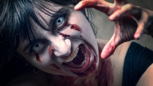 Portrait d'une jeune femme grimée en zombie pour Halloween, avec des lentilles blanches, du faux sang sur le visage et les mains tendues vers l'avant dans une expression effrayante.