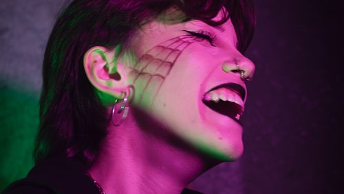 Portrait d'une personne avec un maquillage d'Halloween de toile d'araignée sur le visage, riant avec la bouche ouverte. Elle porte des bijoux argentés, un haut noir et un piercing au nez. L'éclairage est violet et vert.