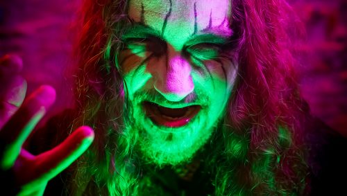 Portrait d'Halloween d'une personne avec des cheveux longs et bouclés, un maquillage vert et blanc sur le visage et un regard menaçant, sous des lumières violettes et vertes.