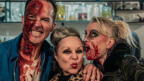 Trois personnes déguisées pour Halloween: l'homme à gauche avec du faux sang sur le visage grimace et montre les dents, la femme au centre fait une moue avec ses lèvres rouges, et celle de droite, avec des faux blessures et du sang sur le visage, a l'air très surprise.