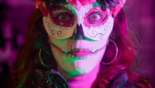 Portrait d'une femme avec un maquillage de squelette coloré et un masque orné de fleurs pour Halloween, éclairée en vert et rose.