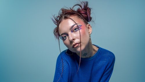 Une jeune femme aux cheveux bruns coiffés en chignon désordonné, avec un maquillage artistique bleu et rose autour des yeux, pose assise, vêtue d'un haut bleu ajouré et d'un pantalon en cuir bleu, portant des bottes noires à semelles épaisses, sur un fond bleu uni, le tout avec un air sérieux et une lumière douce. 