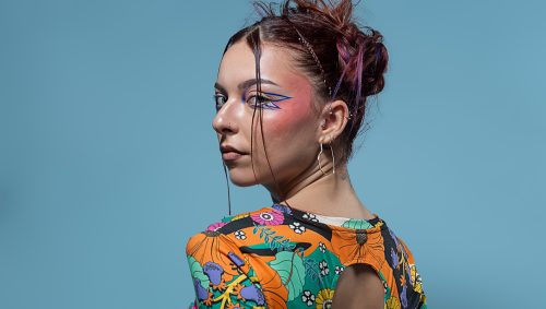 Portrait d'une jeune femme avec un maquillage artistique bleu et rose, portant un haut coloré à motifs floraux et un pantalon bleu en cuir, le tout sur un fond bleu uni. Elle est de dos et regarde vers la gauche, avec une expression neutre.