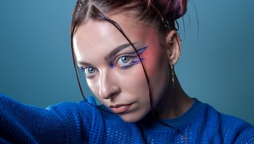 Portrait d'une jeune femme avec un maquillage créatif bleu autour des yeux et des cheveux tressés avec des mèches colorées, regardant directement la caméra sur un fond bleu uni.