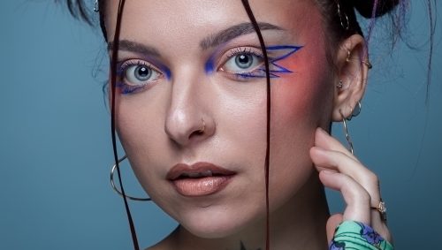 Portrait d'une femme avec un maquillage artistique : fard à paupières bleu et motif graphique sur la tempe, coiffure chignon décoiffé. Elle a l'air calme et confiante. Son haut est imprimé de motifs floraux colorés, et elle porte des boucles d'oreilles créoles. Un tatouage d'étoile orne son cou.