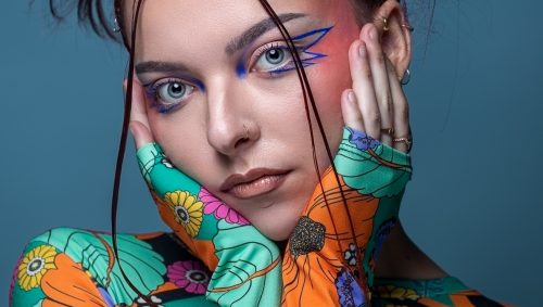 Portrait d'une jeune femme avec un maquillage artistique bleu et rose vif, et une expression neutre, portant un haut fleuri coloré.