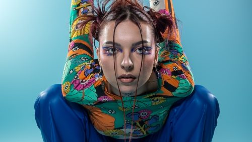 Portrait en studio d'une jeune femme accroupie, regardant fixement l'objectif avec une expression neutre, coiffée de couettes et portant un haut à motifs floraux colorés, un pantalon bleu brillant et des bottes noires à lacets.