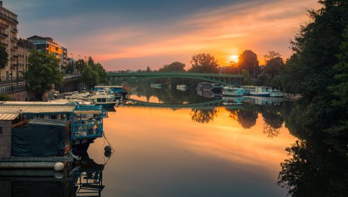 Lever de soleil doré sur Nantes, avec un pont vert qui enjambe la rivière, des bateaux amarrés et des bâtiments qui se reflètent joliment dans l'eau calme. 
