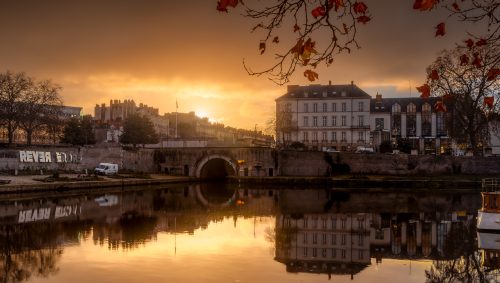 Un lever de soleil doré sur Nantes, avec un canal qui reflète les bâtiments et un pont. Des bateaux sont amarrés au premier plan et des feuilles d'automne encadrent la scène. 
