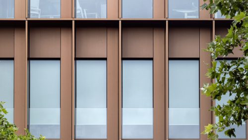 Façade de la Maison de l'Innovation ornée de fenêtres en verre encadrées de métal couleur bronze, avec des arbres verts en partie visibles sur les côtés.
