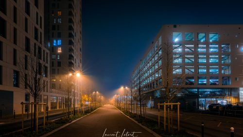 Rue illuminée par des lampadaires avec des bâtiments et un parking couverts une nuit de brouillard à Nantes. 