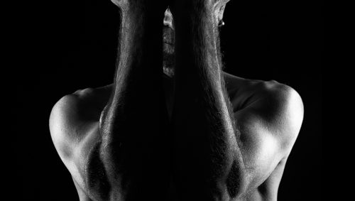 Portrait d'art en noir et blanc d'un homme chauve nu, poings serrés cachant son visage dans l'obscurité.