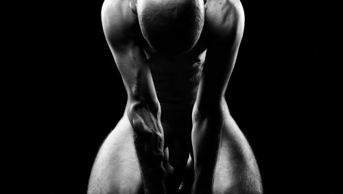 Portrait artistique en noir et blanc d'un homme nu agenouillé, la tête baissée, sur fond noir. 