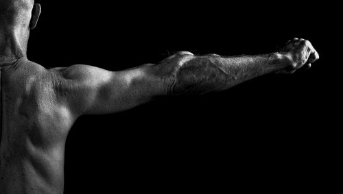 Portrait artistique en noir et blanc d'un homme chauve, vu de dos, bras tendu vers la droite, poing serré, sur fond noir.