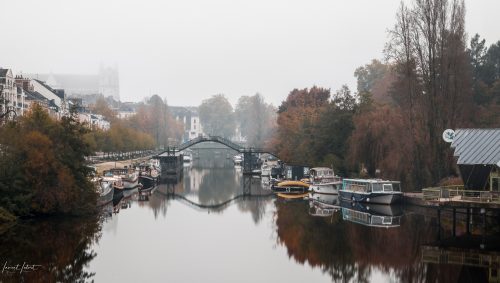 Vue d'automne sur le quai Barbuse et l'île de Versailles à Nantes, enveloppés dans la brume, avec des péniches sur l'eau et la passerelle de l'ile Versailles. En arrière-plan on devinne la cathédrale dans la brume. 
