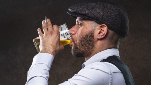Portrait en studio d'un homme à la Peaky Blinders, buvant une bière et fumant une cigarette.