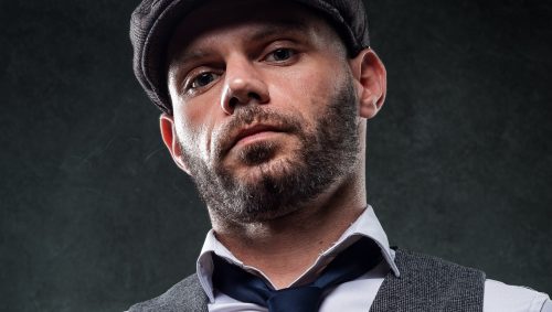 Portrait en contre-plongée d'un homme portant une casquette et un gilet, tenant un verre de bière, avec un regard intense.