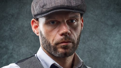 Portrait studio créatif à Nantes : homme barbu avec une casquette à carreaux, un gilet et une cravate dénouée, ambiance Peaky Blinders, regard sérieux.