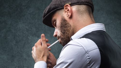 Un homme de profil, avec une casquette de Peaky Blinders, allume une cigarette avec une allumette.
