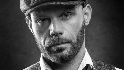 Portrait en noir et blanc d'un homme portant une casquette et un gilet, avec une expression sérieuse.