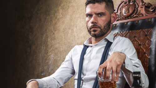 Un homme assis sur un fauteuil en bois sculpté, avec une cigarette à la main et un verre de whisky, une bouteille de Jack Daniel's sur la table d'appoint. 