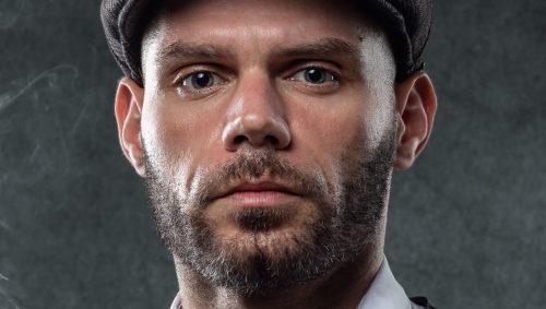 Portrait serré d'un homme avec une casquette en tweed et un regard intense.