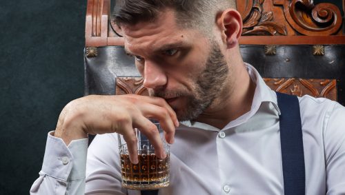 Un homme barbu, avec un regard pensif, est assis sur une chaise en bois sculptée et tient un verre de whisky. Il porte une chemise blanche et des bretelles.
