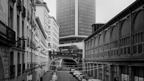 Une rue de Nantes en noir et blanc, avec la Tour de Bretagne à moitié cachée par la brume au fond de l'image. Des bâtiments anciens bordent la rue, et des voitures sont garées le long du trottoir. 